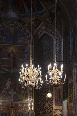 Elaborate chandeliers hang from the ceiling in an ornate setting filled with intricate religious artwork and patterns. The backdrop includes vibrant paintings depicting religious figures and symbols. The atmosphere is richly decorated, giving a sense of historical and cultural significance.