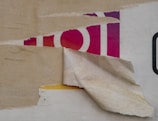 Peeling layers of paper reveal parts of an underlying image, including a portion with red and purple colors and fragments of what appear to be letters or shapes.
