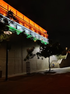 A modern building illuminated with vibrant orange, white, and green lights during nighttime. The building's design features large, glass windows and rows of lights highlighting its structure. The surrounding area includes concrete stairs and a tree casting shadows, with minimal activity.