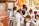 A group of young school children wearing uniforms are gathered near a metal fence, some carrying backpacks. They appear to be waiting or queuing for something. The scene is lively, suggesting a bustling school environment.
