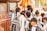 A group of young school children wearing uniforms are gathered near a metal fence, some carrying backpacks. They appear to be waiting or queuing for something. The scene is lively, suggesting a bustling school environment.