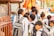 A group of young school children wearing uniforms are gathered near a metal fence, some carrying backpacks. They appear to be waiting or queuing for something. The scene is lively, suggesting a bustling school environment.
