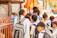 A group of young school children wearing uniforms are gathered near a metal fence, some carrying backpacks. They appear to be waiting or queuing for something. The scene is lively, suggesting a bustling school environment.