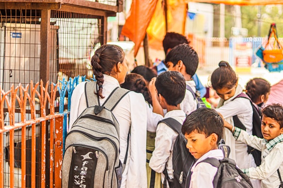 A group of young school children wearing uniforms are gathered near a metal fence, some carrying backpacks. They appear to be waiting or queuing for something. The scene is lively, suggesting a bustling school environment.