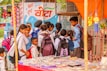 A group of school children wearing uniforms with backpacks stand near a street vendor selling goods under an orange canopy. The background features a colorful mural on a wall with text and images of snacks and drinks on display.
