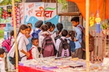 A group of school children wearing uniforms with backpacks stand near a street vendor selling goods under an orange canopy. The background features a colorful mural on a wall with text and images of snacks and drinks on display.