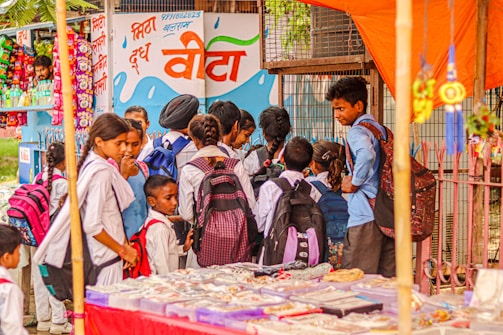 A group of school children wearing uniforms with backpacks stand near a street vendor selling goods under an orange canopy. The background features a colorful mural on a wall with text and images of snacks and drinks on display.