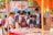A group of school children wearing uniforms with backpacks stand near a street vendor selling goods under an orange canopy. The background features a colorful mural on a wall with text and images of snacks and drinks on display.