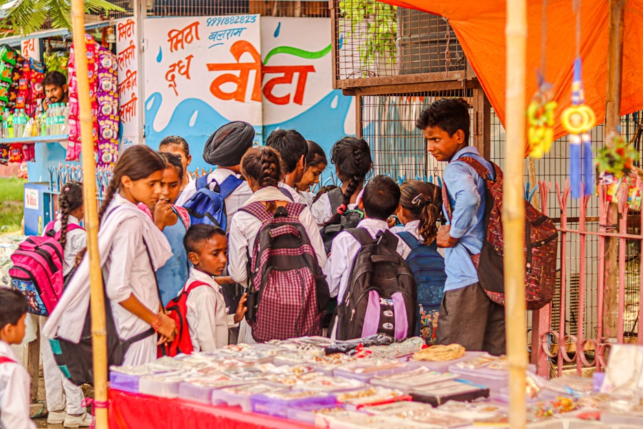A group of school children wearing uniforms with backpacks stand near a street vendor selling goods under an orange canopy. The background features a colorful mural on a wall with text and images of snacks and drinks on display.
