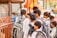 A group of children dressed in school uniforms are gathered closely together at a distance, possibly near a shop or food stall. They carry backpacks and appear to be engaged or interacting with something or someone in front of them.