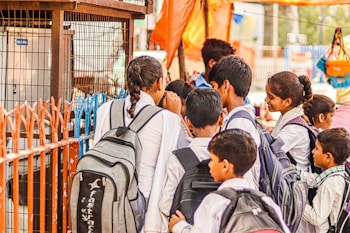 A group of children dressed in school uniforms are gathered closely together at a distance, possibly near a shop or food stall. They carry backpacks and appear to be engaged or interacting with something or someone in front of them.