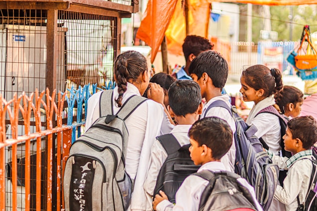 A group of children dressed in school uniforms are gathered closely together at a distance, possibly near a shop or food stall. They carry backpacks and appear to be engaged or interacting with something or someone in front of them.