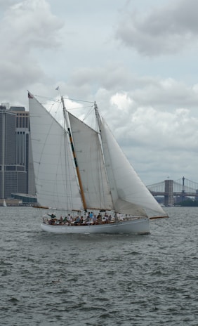 A sailboat with large white sails navigating through a body of water near an urban skyline. The boat is occupied by a group of people, and a bridge is visible in the background, under a cloudy sky.
