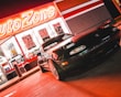A sports car is parked in front of an AutoZone store at night. The store's illuminated sign casts a bright red glow over the scene. The car's sleek design is highlighted by the reflections of the neon lights on its surface. Various promotional posters and products are visible through the store's windows.