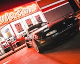 A sports car is parked in front of an AutoZone store at night. The store's illuminated sign casts a bright red glow over the scene. The car's sleek design is highlighted by the reflections of the neon lights on its surface. Various promotional posters and products are visible through the store's windows.