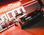 A sports car is parked in front of an AutoZone store at night. The store's illuminated sign casts a bright red glow over the scene. The car's sleek design is highlighted by the reflections of the neon lights on its surface. Various promotional posters and products are visible through the store's windows.