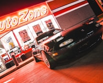 A sports car is parked in front of an AutoZone store at night. The store's illuminated sign casts a bright red glow over the scene. The car's sleek design is highlighted by the reflections of the neon lights on its surface. Various promotional posters and products are visible through the store's windows.