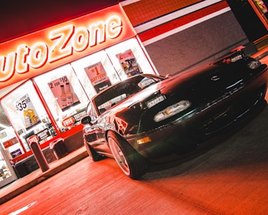 A sports car is parked in front of an AutoZone store at night. The store's illuminated sign casts a bright red glow over the scene. The car's sleek design is highlighted by the reflections of the neon lights on its surface. Various promotional posters and products are visible through the store's windows.