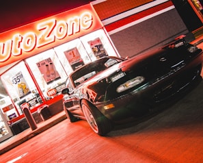 A sports car is parked in front of an AutoZone store at night. The store's illuminated sign casts a bright red glow over the scene. The car's sleek design is highlighted by the reflections of the neon lights on its surface. Various promotional posters and products are visible through the store's windows.