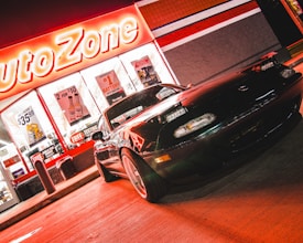 A sports car is parked in front of an AutoZone store at night. The store's illuminated sign casts a bright red glow over the scene. The car's sleek design is highlighted by the reflections of the neon lights on its surface. Various promotional posters and products are visible through the store's windows.