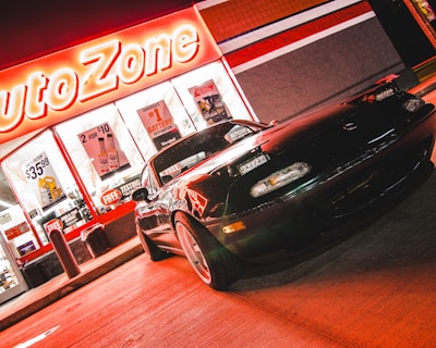 A sports car is parked in front of an AutoZone store at night. The store's illuminated sign casts a bright red glow over the scene. The car's sleek design is highlighted by the reflections of the neon lights on its surface. Various promotional posters and products are visible through the store's windows.