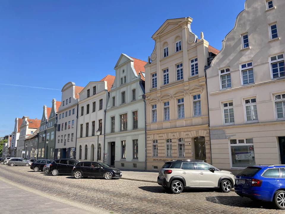 The image depicts a row of old, historic buildings lining a cobblestone street in a European city. The buildings feature ornate architectural details and are painted white with red roofs, creating a picturesque scene. Cars are parked along the side of the road, adding to the urban feel of the setting. The clear blue sky suggests pleasant weather conditions for the day.