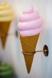 A pink swirl of soft serve ice cream is neatly placed in a waffle cone, which is held up by a cone holder attached to a wall.