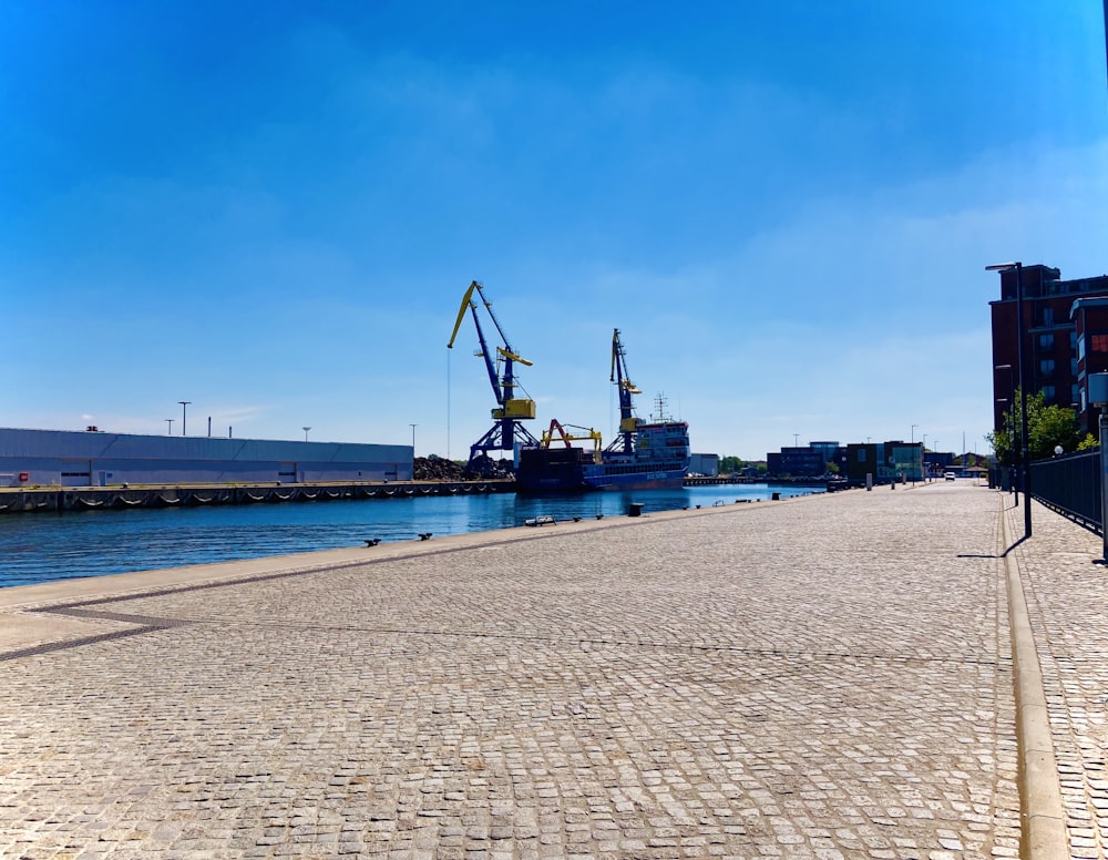 a brick walkway next to a body of water with cranes in the background