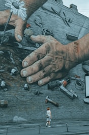 A large mural painted on a brick wall depicting two human hands working with mechanical tools and a white daisy. Below the mural, a person with red hair is walking on the sidewalk, adding a sense of scale.