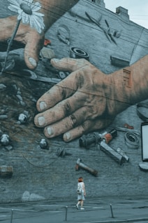 A large mural painted on a brick wall depicting two human hands working with mechanical tools and a white daisy. Below the mural, a person with red hair is walking on the sidewalk, adding a sense of scale.