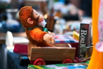 A plush monkey toy is seated in a small wooden toy cart with red wheels. The surrounding area is cluttered with various items, including a board game box labeled 'Roulette' and a book with colorful illustrations.