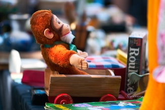 A plush monkey toy is seated in a small wooden toy cart with red wheels. The surrounding area is cluttered with various items, including a board game box labeled 'Roulette' and a book with colorful illustrations.