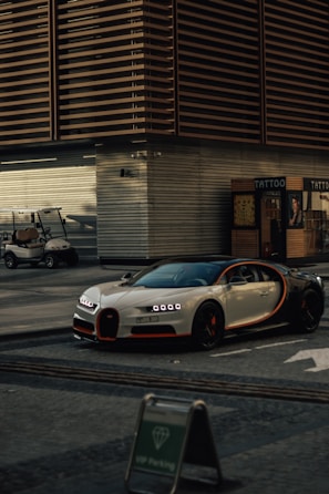 A luxury sports car with a sleek design and distinctive headlights is parked on a street. It is positioned near a modern building with horizontal paneling and next to a shop with a sign that reads 'TATTOO'. A small golf cart is also visible in the background, and a sign indicating VIP parking is placed in front of the car.