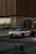 A luxury sports car with a sleek design and distinctive headlights is parked on a street. It is positioned near a modern building with horizontal paneling and next to a shop with a sign that reads 'TATTOO'. A small golf cart is also visible in the background, and a sign indicating VIP parking is placed in front of the car.