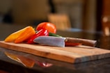 A wooden cutting board holds a variety of colorful bell peppers and a knife. The peppers include yellow, red, and orange varieties, paired with a metal-bladed knife with a wooden handle. The background is blurred, suggesting a kitchen environment.