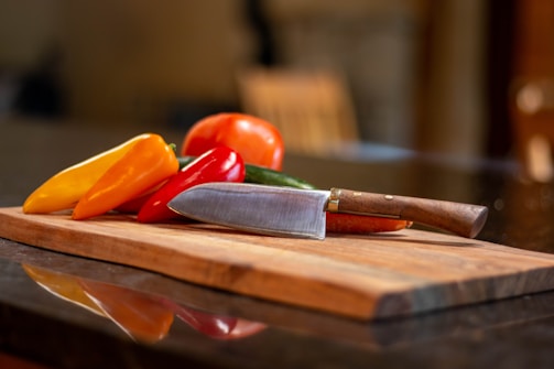 A wooden cutting board holds a variety of colorful bell peppers and a knife. The peppers include yellow, red, and orange varieties, paired with a metal-bladed knife with a wooden handle. The background is blurred, suggesting a kitchen environment.