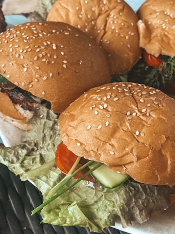 Four freshly prepared burgers with sesame seed buns are placed on a surface. Each burger contains a variety of fresh vegetables including lettuce, tomato, cucumber, and possibly herbs. The buns are golden brown, and the overall presentation is appetizing.