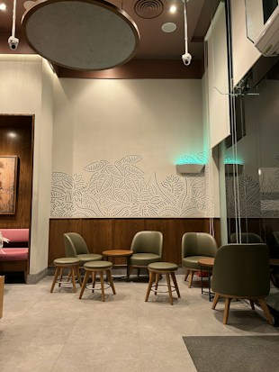 A cozy indoor seating area with green cushioned chairs and wooden stools arranged neatly around wooden tables. The walls are adorned with decorative leaf patterns and a mix of light and dark wooden panels. Soft lighting complements the room's warm tone, with ceiling fixtures visible.