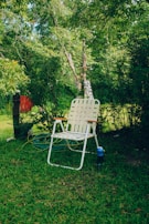 A white woven lawn chair with wooden armrests is placed on lush green grass. Behind the chair, a garden hose is coiled on the ground. The background is filled with dense green foliage and trees, creating a tranquil and natural setting.