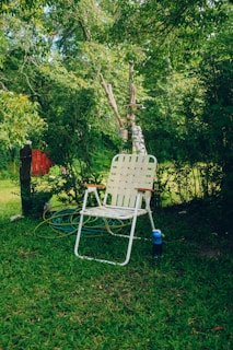 A white woven lawn chair with wooden armrests is placed on lush green grass. Behind the chair, a garden hose is coiled on the ground. The background is filled with dense green foliage and trees, creating a tranquil and natural setting.