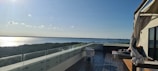 A rooftop terrace with a modern glass railing overlooks a calm body of water and a distant treeline. The wooden flooring is accompanied by a few pieces of outdoor furniture, including a chair and a daybed with cushions. A canopy with flowing drapes adds an elegant touch to the setting. The sunlight reflects off the water, adding brightness to the serene view.