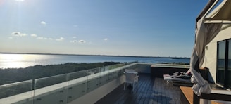 A rooftop terrace with a modern glass railing overlooks a calm body of water and a distant treeline. The wooden flooring is accompanied by a few pieces of outdoor furniture, including a chair and a daybed with cushions. A canopy with flowing drapes adds an elegant touch to the setting. The sunlight reflects off the water, adding brightness to the serene view.