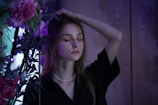 A young woman stands with her eyes closed, her right hand resting on her head. She wears a black top and a pearl necklace. Around her are vibrant flowers and green leaves, under dim, moody lighting that casts a purple hue.