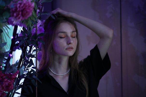 A young woman stands with her eyes closed, her right hand resting on her head. She wears a black top and a pearl necklace. Around her are vibrant flowers and green leaves, under dim, moody lighting that casts a purple hue.