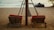 Two wooden lounge chairs with red cushions are positioned on a sandy beach, facing towards the ocean. A wooden structure with a woven canopy stands between the chairs. In the background, the calm sea and sky meet at the horizon, and there are a few indistinct figures in the water.