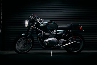 A sleek, dark-colored motorcycle with chrome accents is parked against a backdrop of horizontal metal shutters. The motorcycle has a classic cafe racer design with a powerful engine, round headlight, and shiny exhaust pipes. The word 'Thruxton' is visible on its body.