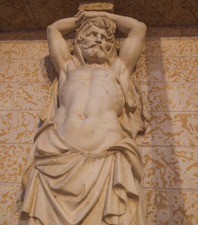 A classical stone sculpture of a muscular, bearded man with a draped cloth around his waist, posed in a manner reminiscent of ancient Roman or Greek statues. The background consists of a textured wall.