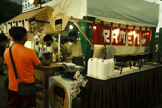 A food stall in a market is depicted, with a prominent sign displaying 'RAMEN'. Several people are gathered, some serving and some ordering. Various cooking utensils and ingredients, including bottles and grills, are visible.