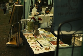 A market stall displays an array of vibrant greeting cards and floral arrangements. Next to the cards, a decorative sign and small ornaments are placed on the table. In the background, people browse through other stalls, creating a lively and community-focused atmosphere.