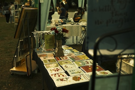 A market stall displays an array of vibrant greeting cards and floral arrangements. Next to the cards, a decorative sign and small ornaments are placed on the table. In the background, people browse through other stalls, creating a lively and community-focused atmosphere.